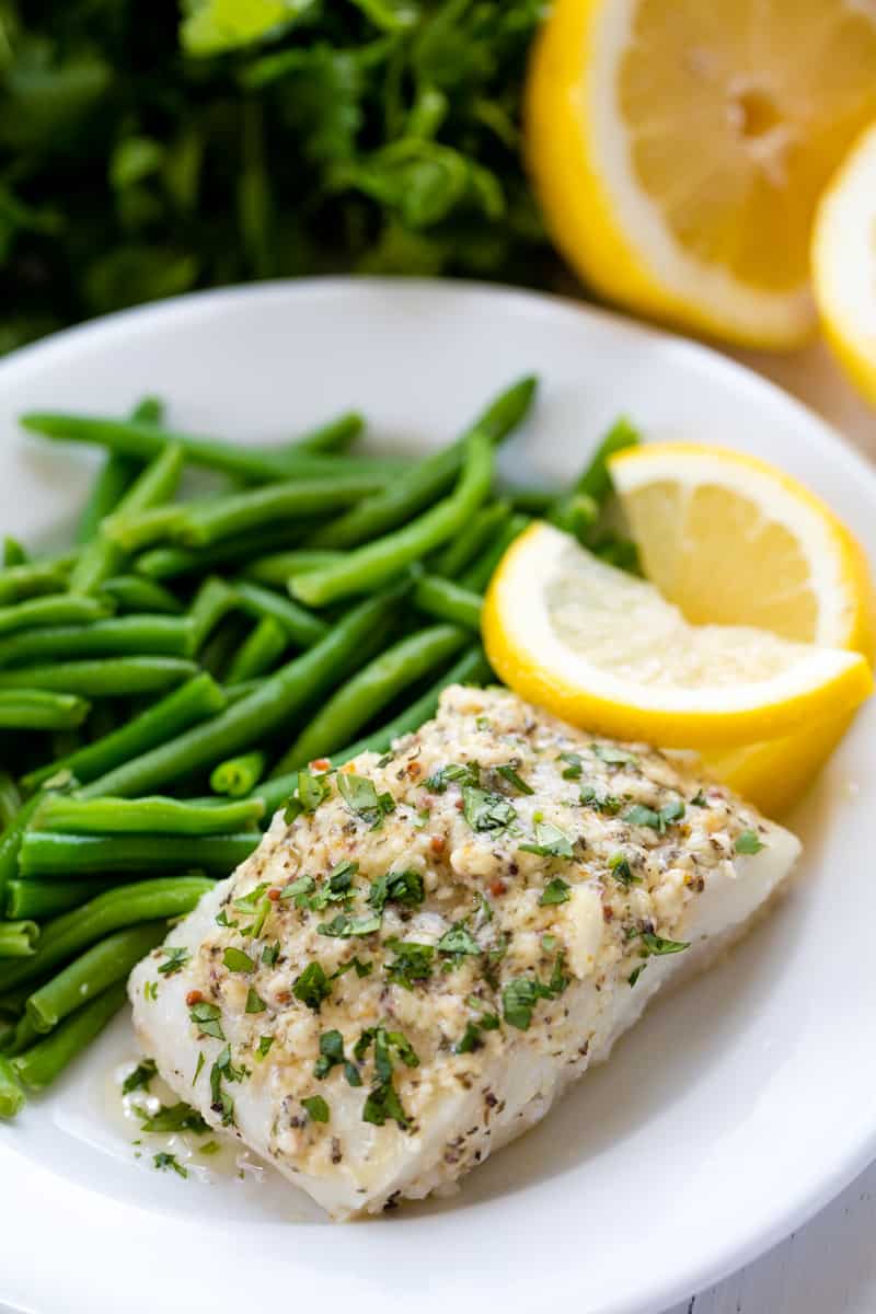 Lemon baked codfish served up on a white plate with lemon slices and green beans.