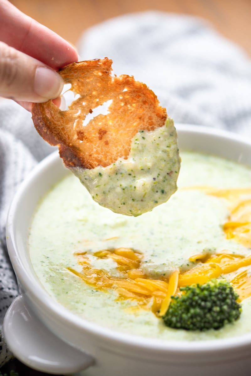 A slice of crusty toasted baguette being dipped into a bowl of cream of broccoli soup.