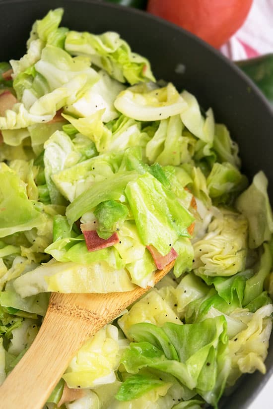 Cut cabbage and bacon bits in a pan with a wooden spoon in it.