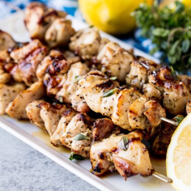 A stack of Grilled Lemon and Herb Chicken Thigh Skewers on a white serving plate with a half lemon.